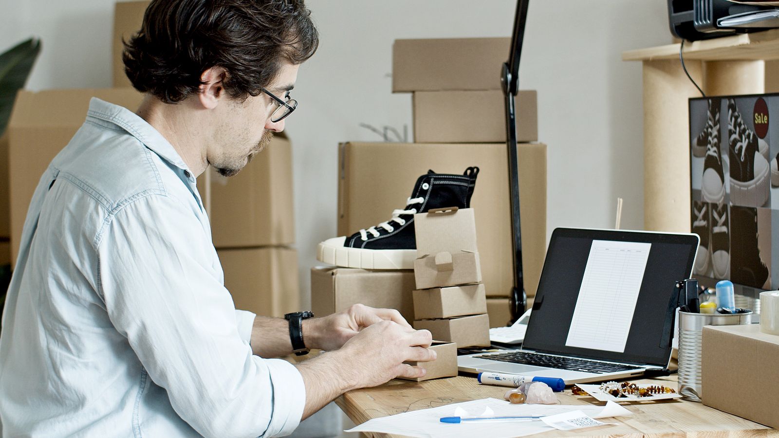 Commerçant en ligne préparant les commandes de sa boutique, entouré de cartons, de produits et d'un ordinateur portable.
