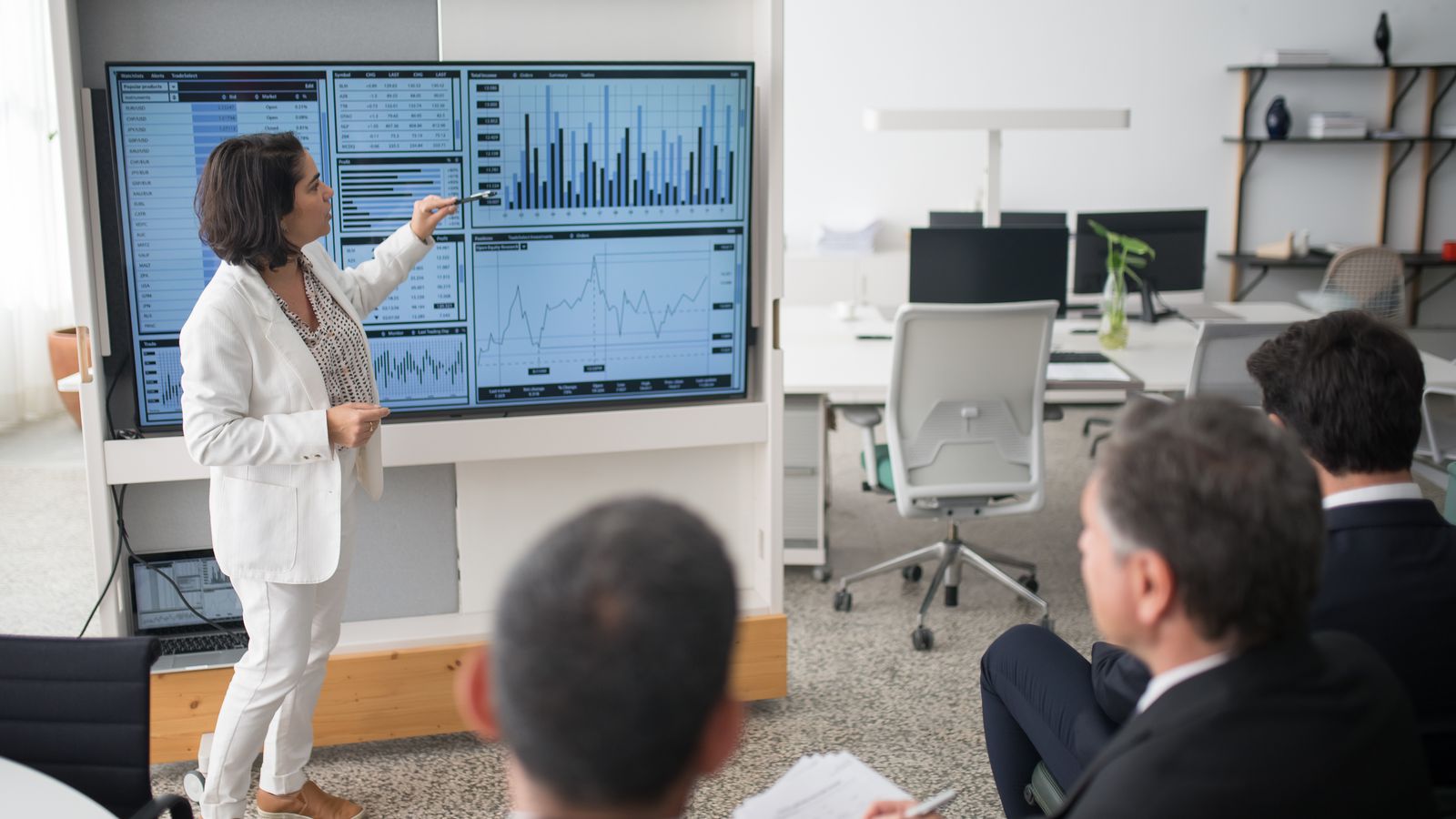 Consultante présentant un tableau de bord de données à des dirigeants en costume assis dans une salle de réunion.